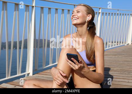 Glückliche Frau, die Musik über kabellose in-Ear-Kopfhörer hört, die ihr Mobiltelefon auf einem erhöhten Gehweg halten Stockfoto