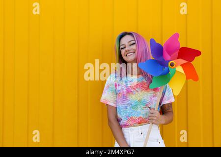 Fröhliche Frau, die ein mehrfarbiges Kleinrad vor der gelben Wand hält Stockfoto