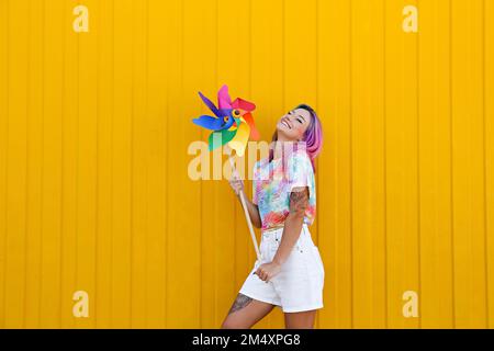 Lächelnde Frau, die ein mehrfarbiges Kleinrad vor der gelben Wand hält Stockfoto