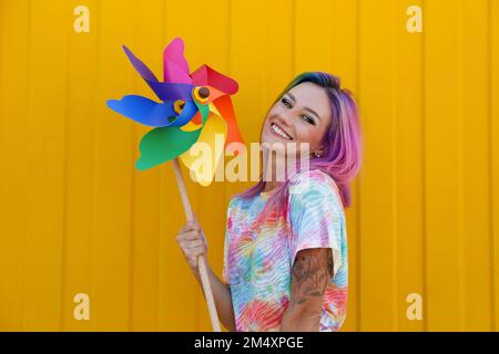 Fröhliche junge Frau, die ein mehrfarbiges Kleinrad vor der gelben Wand hält Stockfoto