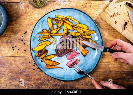 Beschreibung: Ein Mann hält ein hausgemachtes Filetsteak mit knusprigen goldbraunen Kartoffelspalten auf einem blauen Teller neben einem Steinmort Stockfoto