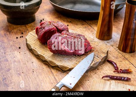 Beschreibung: Zubereitung von rohem Rinderfiletsteak auf Holzbrett mit Stahlpfanne, Mörtelsalz und Pfeffershakerand Rosmarin und kühlem Schneidebrett p Stockfoto