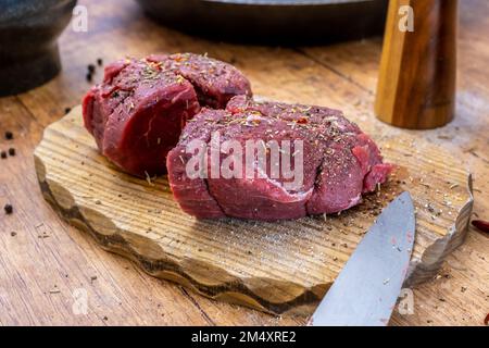 Beschreibung: Zubereitung von rohem Rinderfiletsteak auf Holzbrett mit Stahlpfanne, Mörtelsalz und Pfeffershakerand Rosmarin und kühlem Foto von fr Stockfoto