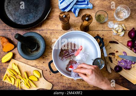 Beschreibung: Zubereitung von rohem Rinderfiletsteak für ein Sous-Vide-Bad in einem Topf mit Thermometer auf einem Holztisch mit Stahlpfanne, Mörtel und Küchentuch Stockfoto