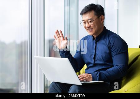 Ein junger asiatischer Student sitzt auf dem Campus, arbeitet zusammen und lernt online von einem Laptop aus. Er spricht durch einen Videoanruf, grüßt mit der Hand, lächelt. Stockfoto