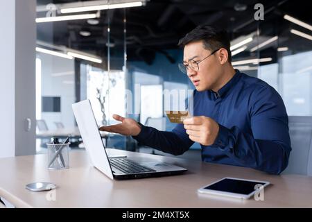 Besorgter junger Asiate, der im Büro am Tisch sitzt und eine Kreditkarte hält. Er sah schockiert auf dem Laptop-Monitor aus und spreizte seine Hände. Scheckkonto, fehlgeschlagene Online-Käufe, Konkurs. Stockfoto