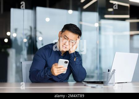 Porträt eines besorgten jungen Asiaten, der im Büro am Tisch sitzt, seinen Kopf mit der Hand hält und traurig auf das Telefon schaut. Gelangweilt, schlechte Nachrichten, Nachrichten. Stockfoto