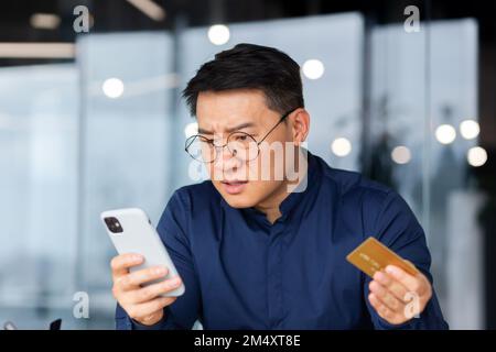 Nahaufnahme. Ein besorgter junger Asiate mit Brille hat eine Kreditkarte und sieht schockiert vom Telefon aus. Scheckkonto, fehlgeschlagene Online-Käufe, Konkurs. Stockfoto