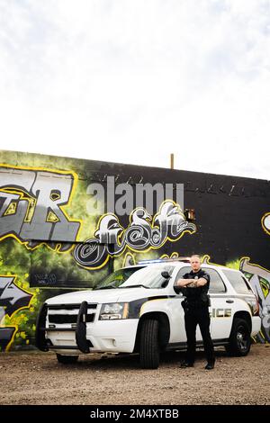 Vertikales Bild eines weißen, männlichen, weißen Polizisten mit gekreuzten Armen, der vor seinem Auto steht und Graffiti trägt Stockfoto