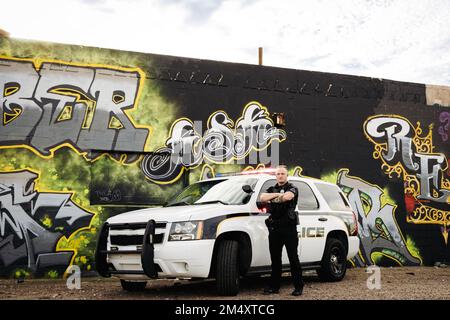 Horizontales Bild eines weißen, männlichen, weißen Polizisten mit gekreuzten Armen, der vor seinem Auto mit Graffiti in seiner Stadt steht Stockfoto