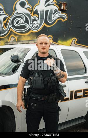 Vertikales Bild eines weißen, männlichen, weißen Polizisten, der mit geradem Gesicht posiert und eine Hand auf einer kugelsicheren Weste, die vor seinem Polizeiauto steht Stockfoto