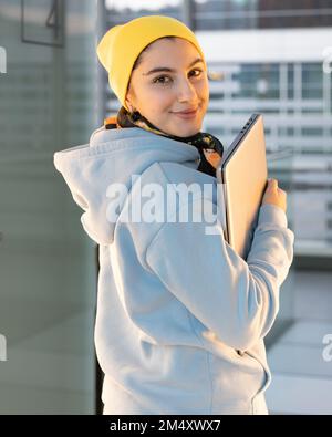 Lächelnde Studentin in blauem Sweatshirt und gelbem Hut läuft mit einem Computer den Flur entlang Stockfoto