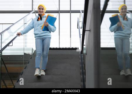 Eine lächelnde Studentin mit bunten Ordnern in der Hand geht die Treppe hinunter Stockfoto