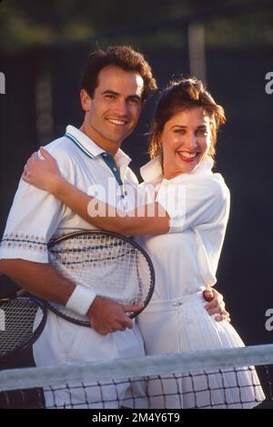 Ein junges weißes Paar in Tennisbekleidung posiert vor dem Netz, umarmt sich und lächelt in die Kamera Stockfoto