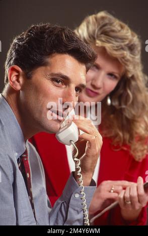 Bürosituation mit einem Geschäftsmann am Telefon, während eine Sekretärin im Hintergrund Notizen macht Stockfoto