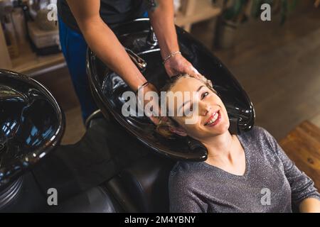 Blick aus dem Blickwinkel auf eine lächelnde, entspannte Frau, deren Haare von einem unbekannten Friseur gewaschen und massiert werden. Haare im Friseursalon waschen. Hochwertiges Foto Stockfoto