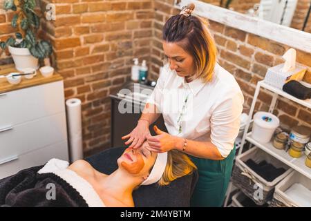 Gesichtsmassage aus dem hohen Blickwinkel. Positive weisse Frau, die auf DEM SPA-Bett liegt, während eines Gesichtseingriffs durch einen Kaukasier-Spezialisten. Hochwertiges Foto Stockfoto