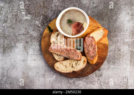 Hausgemachte Hühnerleber-Pastete auf knusprigem Brot auf einem runden Holzschneidebrett auf dunkelgrauem Hintergrund. Draufsicht, flach liegend Stockfoto