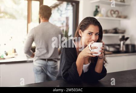 Kaffee macht den MMM morgens. Porträt einer jungen Frau, die in der Küche Kaffee trinkt, während ihr Mann im Hintergrund steht. Stockfoto