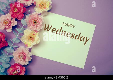 „Happy Wednesday“-Schriftzug und Blumendekoration auf lila Hintergrund Stockfoto