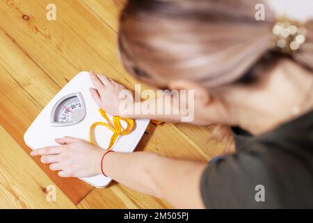 Eine traurige Frau mit Maßband sieht eine weiße Skala auf einem Holzboden Stockfoto