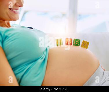 Demnächst verfügbar. Eine schwangere Frau, die zu Hause mit Babyblöcken aus Holz auf dem Bauch liegt. Stockfoto