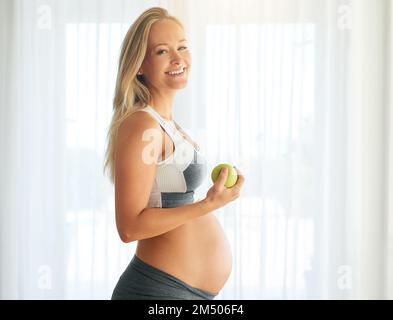 Gesunde Entscheidungen für mein Baby treffen. Porträt einer glücklichen schwangeren Frau in Sportkleidung und Apfelessen zu Hause. Stockfoto