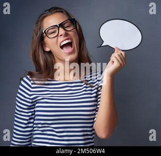 Geben Sie Ihrer Meinung eine Stimme. Studiofoto einer attraktiven jungen Frau, die eine leere Sprechblase hält und vor grauem Hintergrund schreit. Stockfoto