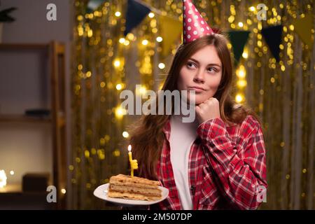 Eine denkende junge Frau mit Partyhut, die mit der Hand nach oben auf dem Gesicht schaut und ein Stück Kuchen hält, während sie zu Hause eine Geburtstagsparty feiert. Verstanden Stockfoto