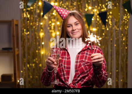 Fröhliche junge Frau mit Konus-Partyhut mit leuchtenden Glitzern und Champagner in den Händen während der Geburtstagsfeier zu Hause. Speicherplatz kopieren Stockfoto