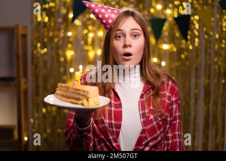 Eine überraschte junge Frau mit einem Hut auf dem Kopf feiert Geburtstag unter Freunden, verblüfft von einem unerwarteten Geschenk. Speicherplatz kopieren Stockfoto