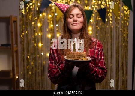 Ein glückliches 18-jähriges Mädchen in Partykegel wünscht sich etwas und bläst Kerzen zum Geburtstagskuchen aus, feiert zu Hause Geburtstag. Speicherplatz kopieren Stockfoto