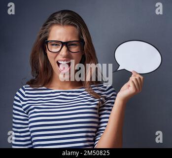 Ich werde nicht ruhig bleiben, ich werde es ausrufen. Studiofoto einer attraktiven jungen Frau, die eine leere Sprechblase hält und vor grauem Hintergrund schreit. Stockfoto