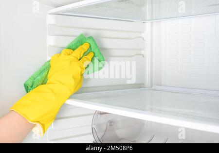 Tiefenreinigung. Hausreinigung. Waschkühlschrank. Weibliche Hand in gelbem Gummischutzhandschuh, der weiß, offen, leer, Kühlschrank mit Grün reinigt Stockfoto