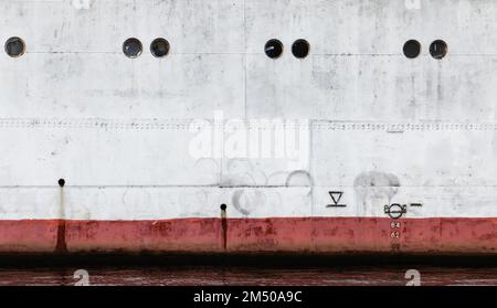 Klassischer weißer Schiffsrumpf mit roter Wasserlinie und Bullaugen, industrielle Hintergrundstruktur Stockfoto