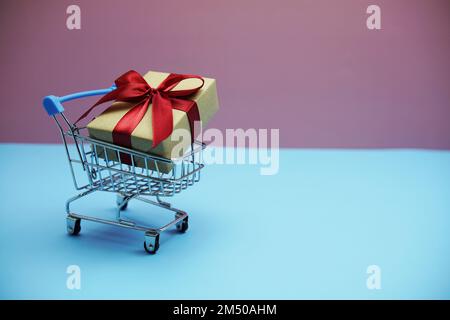 Geschenkbox mit rotem Band im Einkaufswagen auf pinkfarbenem und blauem Hintergrund Stockfoto