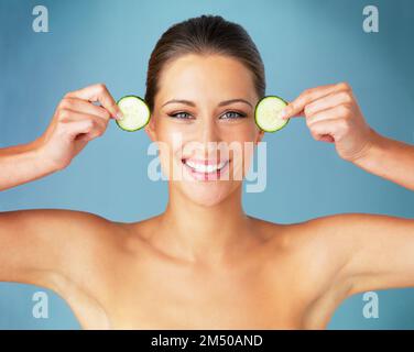Meine Haut liebt Bio. Studio-Porträt einer wunderschönen jungen Frau, die Gurkenschnitte bis ins Gesicht hält, vor blauem Hintergrund. Stockfoto