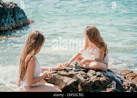 Eine Frau mit Tochter hat eine Biltsteinpyramide am Meer an einem sonnigen Tag auf blauem Meereshintergrund. Frohe Familienferien. Kieselstrand, ruhiges Meer. Konzept Stockfoto