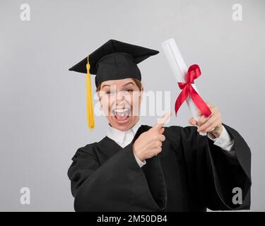 Glückliche Frau im Graduiertenkleid, die mit dem Finger auf ein Diplom zeigt. Stockfoto