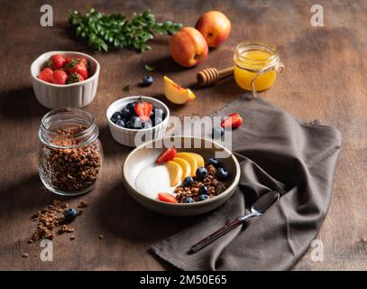 Konzept eines gesunden und nahrhaften Frühstücks. Köstlicher Naturjoghurt mit hausgemachtem Müsli, Pfirsich, Erdbeere, Honig und Heidelbeere in einer Schüssel auf einem Stockfoto