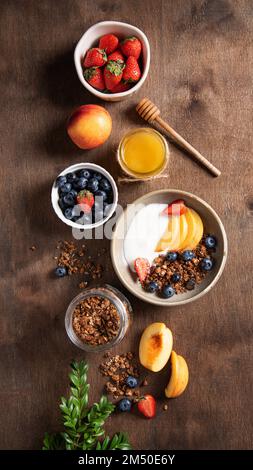 Flach liegender köstlicher natürlicher Joghurt mit hausgemachtem Müsli, Pfirsich, Honig und Beeren in einer Schüssel auf dunklem Holzhintergrund. Das Konzept eines gesunden und Stockfoto