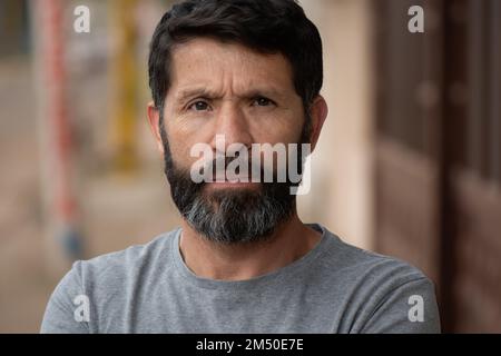Eine Nahaufnahme eines bärtigen, schwarzhaarigen Mannes, der ein graues Hemd trägt, mit einem ernsthaften Blick, der Falten auf seinem Gesicht mit Stirnrunzeln hervorhebt Stockfoto