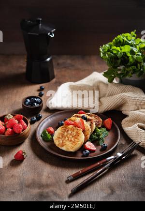 Hüttenkäse-Pfannkuchen oder Syrniki mit Minze, Erdbeeren und Blaubeeren auf einem dunklen Holztisch mit einer schwarzen Kaffeekanne. Konzept dunkel und launisch Stockfoto