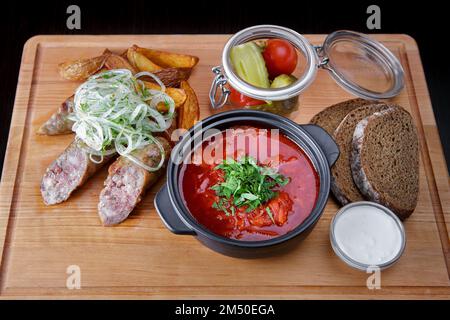 Ukrainischer Borschtsch mit Sauerrahm, Scheiben von gebratenen Kartoffeln und gebratener Wurst auf einem Holzbrett Stockfoto