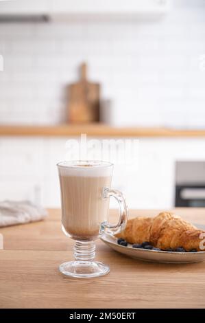 Ein Glas duftender Cappuccino-Kaffee mit Zimt und Croissant auf einem Holztisch vor dem Hintergrund einer weißen Küche am frühen Morgen. Bre Stockfoto