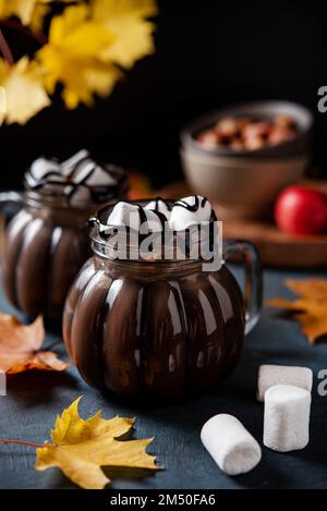 Zwei Tassen aromatische heiße Schokolade mit Marshmallows auf dunklem Hintergrund mit Ahornblättern im Herbst. Konzept gemütlicher Drink. Vorderansicht und Nahaufnahme. Stockfoto