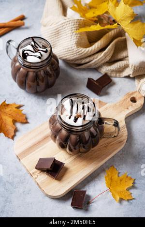 Zwei Tassen heiße Schokolade mit Marshmallows und Zimt auf blauem Hintergrund mit Ahornblättern im Herbst. Konzept gemütlicher Drink. Draufsicht. Stockfoto