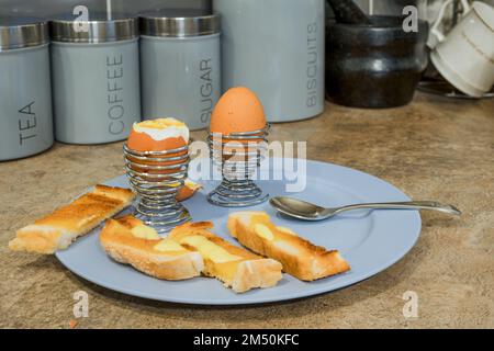 Zwei gekochte Eier in Eierbechern aus Chromdraht mit getoasteten Brotsoldaten und Butterlaschen Stockfoto