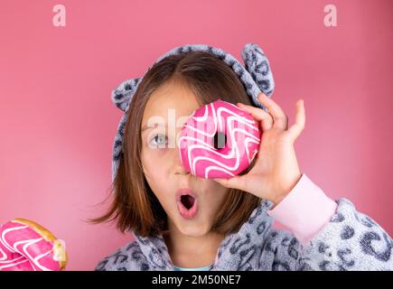 Mädchen in Kigurumi oder Pyjama mit Donuts anstelle des Auges auf pinkfarbenem Hintergrund. Hochwertiges Foto Stockfoto