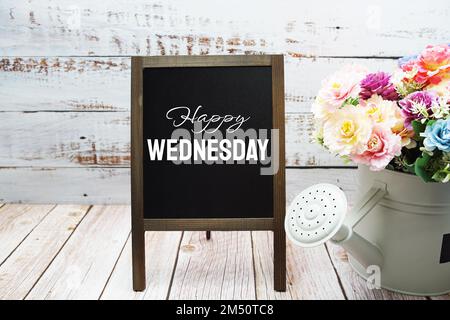 „Happy Wednesday“-Text an der Tafel mit Blumenstrauß auf Holzhintergrund Stockfoto